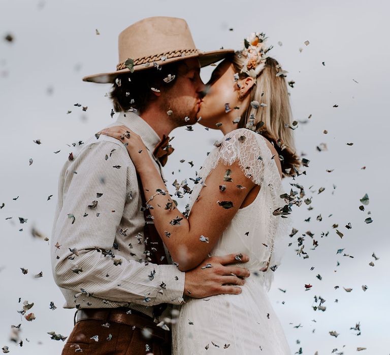 Boho wedding confetti moment with the groom wearing a stone coloured hat and chain kissing the bride in a lace wedding dress and flower crown