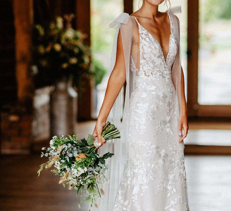 The bride wears a fitted Made With Love lace wedding dress with tulle wings and bow straps holding a neutral wedding bouquet for rustic luxe style wedding