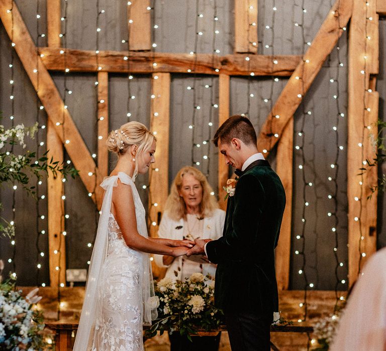 Groom in green velvet tuxedo standing at the altar with the bride in a lace wedding dress with tulle wings standing in front of fairy light backdrop for rustic luxe wedding 