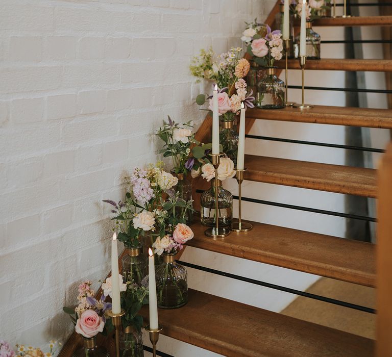 Pastel wedding flowers in shades of pink, purple, cream, and orange with white taper candles in gold holders decorating the stairs 