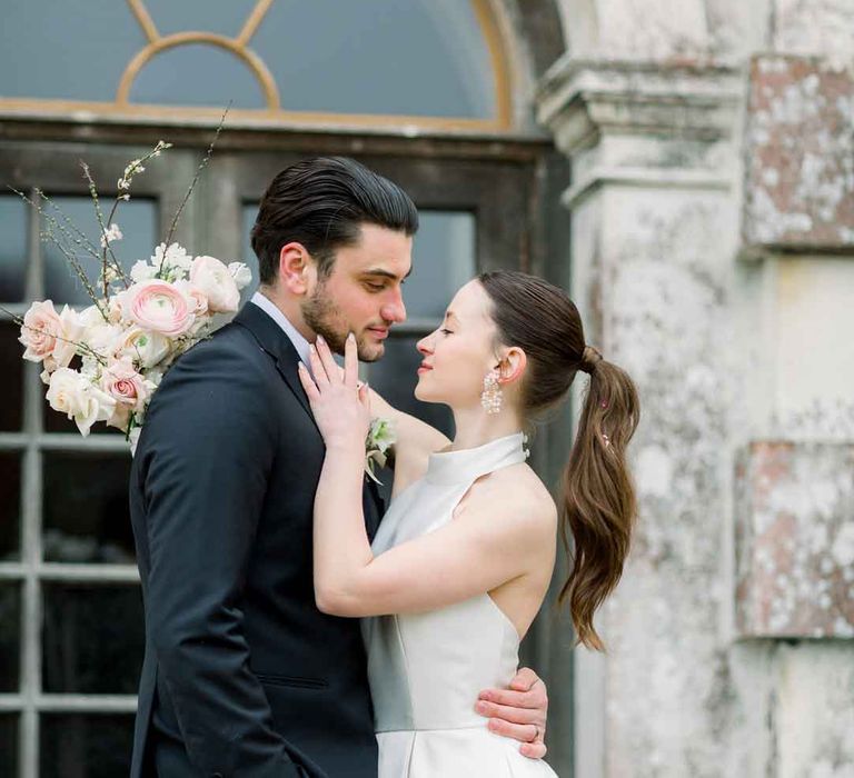 Groom in classic black tuxedo and bride in satin look halterneck wedding dress embracing at St Giles House 