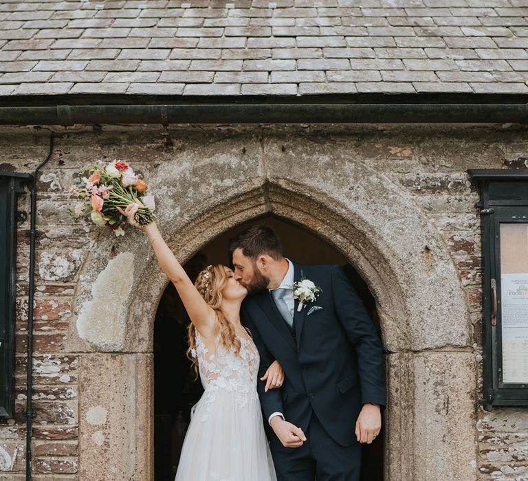 Bride in lace wedding dress raising her pastel wedding bouquet kissing the groom in navy suit after wedding 