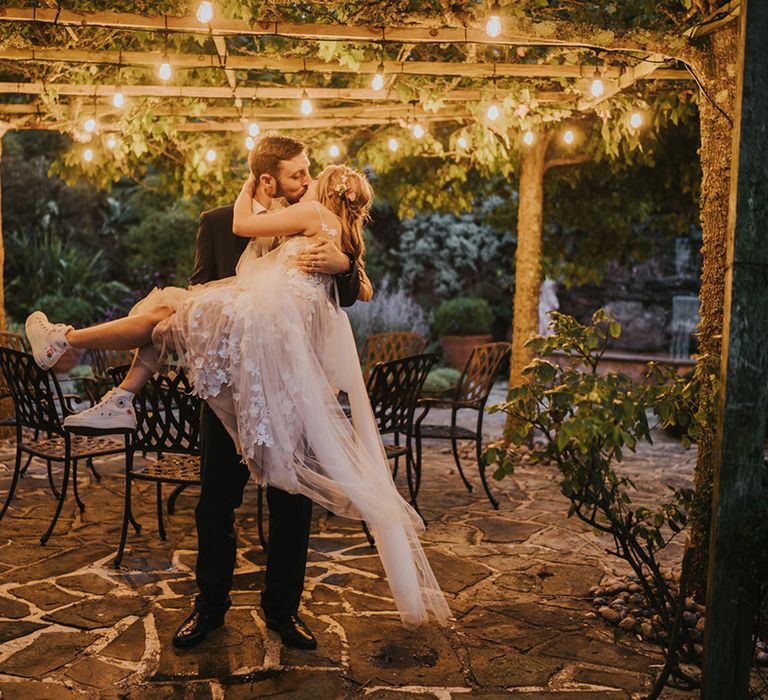 Groom lifts the bride up to kiss her as she wears a tulle Anna Kara wedding dress and embroidered custom Converse trainers 