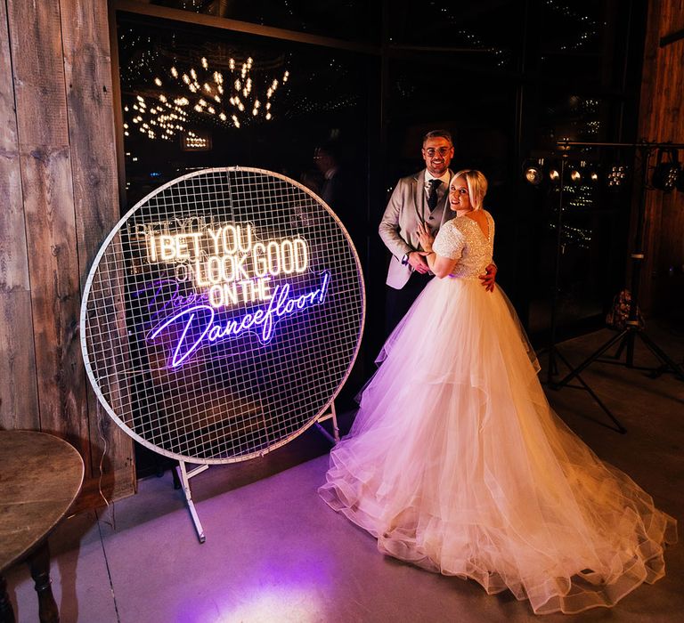 The bride and groom pose next to a decorative neon wedding sign with white and purple neon colours