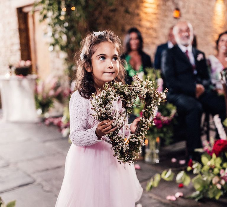 Flower girl in pink tulle dress with knitted cardigan holding a heart shaped floral arrangement walking down the aisle 