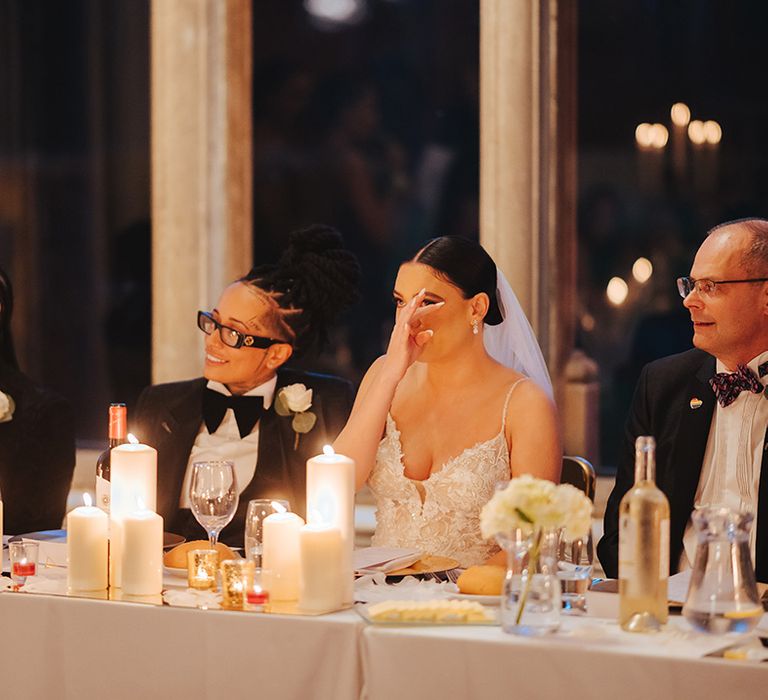 Bride wearing lace sleeveless v neck Enzoani wedding dress wiping tear from her eye sitting next to bride in black snakeskin print tuxedo with black bowtie and white rose boutonniere at head table of Grittleton House