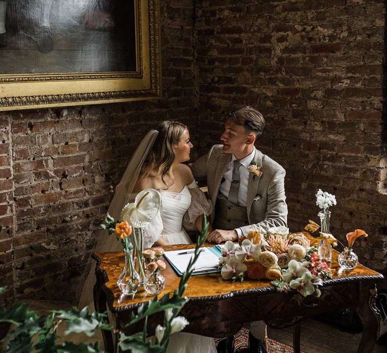 The bride and groom sit to sign their wedding register with seasonal floral arrangements