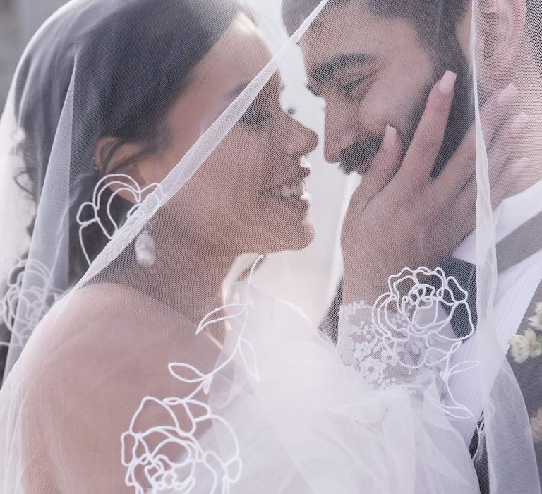 Bride in off the shoulder wedding dress with detachable puff tulle sleeves and groom in classic black tux embracing underneath personalised embroidered wedding veil at Pentney Abbey wedding