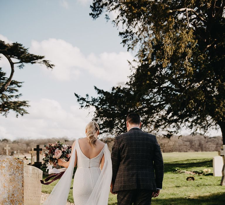 Bride in elegant floral lace fitted wedding dress with tulle wings walking with the groom in Marc Dary grey wedding suit 