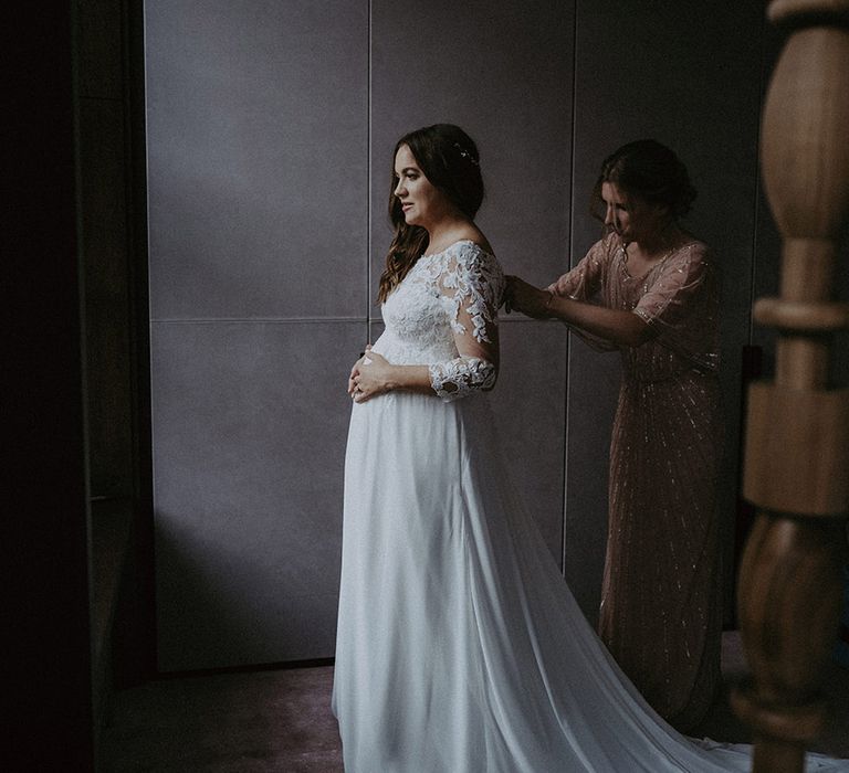 Bride in lace Pronovias maternity wedding dress with her curled brown hair being buttoned up by a wedding guest in sparkly dress 