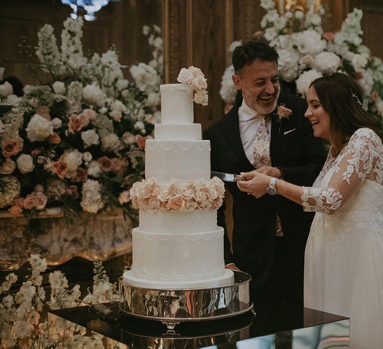 The pregnant bride in a lace wedding dress with groom cutting the six tier luxe wedding cake 