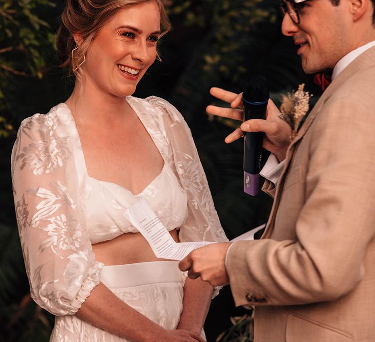 Groom in cream suit reads out his personalised wedding vows with the bride in lace separates smiling brightly at the groom with gold earrings 