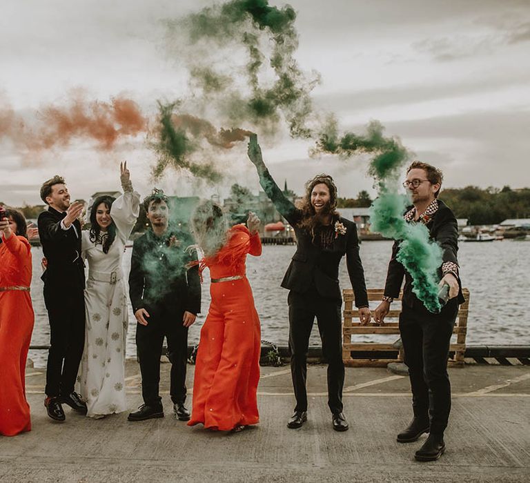 Bridesmaids in orange Bowen Dryden outfits waving colourful smoke bombs with the rest of the wedding party celebrating the retro wedding 