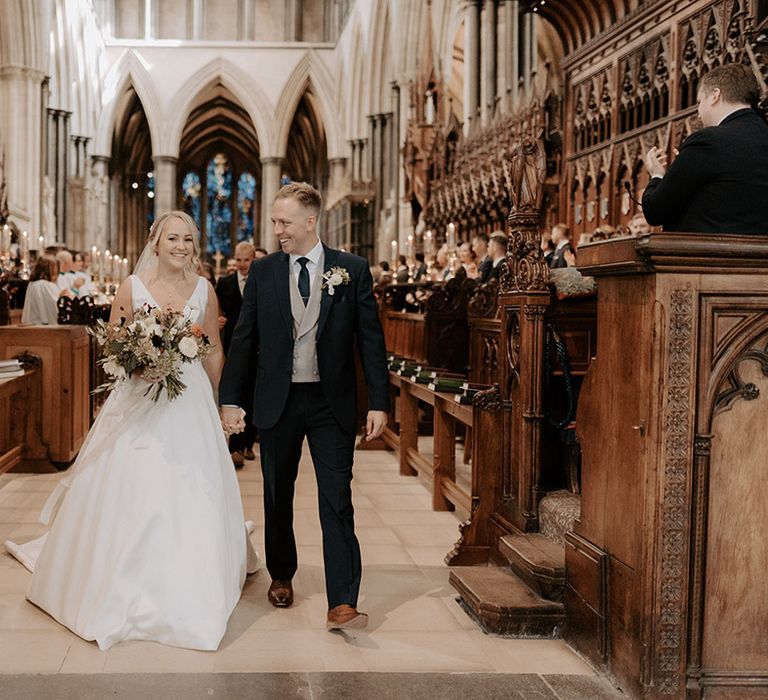 The bride in a plunging wedding dress walks back down the aisle with the groom in a navy suit as a married couple 