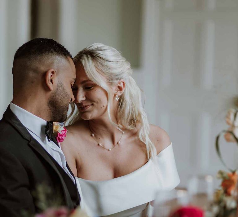 Groom in classic three piece grooms tuxedo with colourful spring boutonniere and bride in off the shoulder lace wedding dress sat at spring wedding tablescape at Frensham Hall wedding venue 