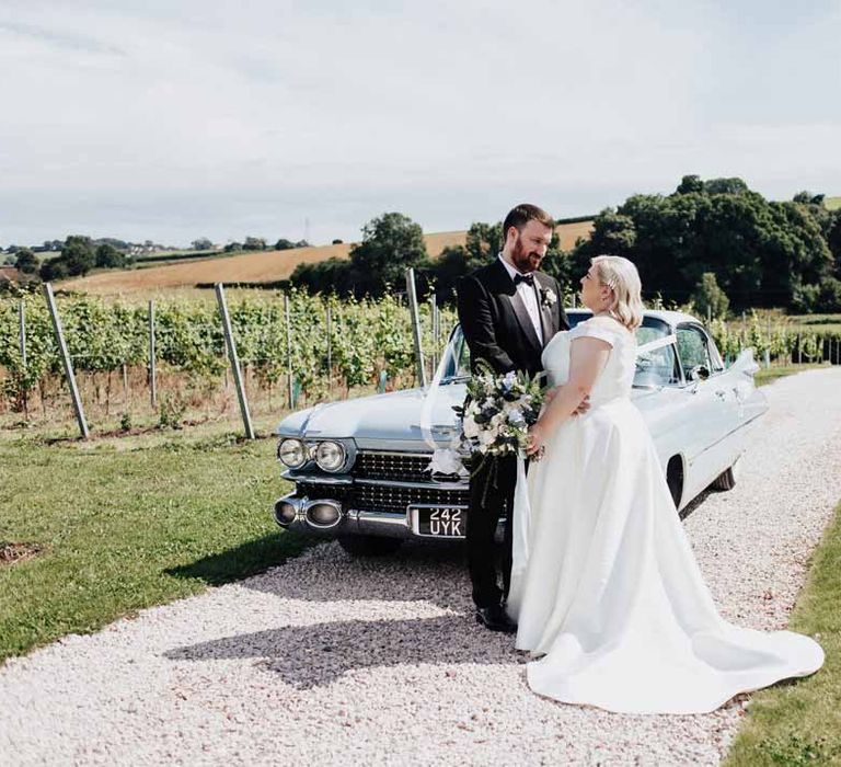 Groom in classic black grooms three piece tuxedo with white rose boutonniere and black bowtie embracing bride in satin off the shoulder wedding dress holding white and blue eucalyptus, foliage, garden rose, gardenia, light blue delphinium and dried flower bridal bouquet tied with dusky blue ribbon standing beside baby blue classic wedding car 