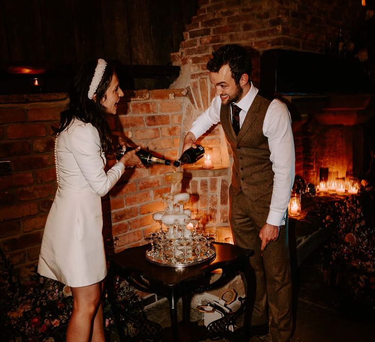 Bride in long sleeve short wedding dress, pearl bridal headband and platform pearl wedding heels standing with groom in brown waistcoat and suit trousers doing wedding champagne tower at Willow Marsh Farm Loughborough