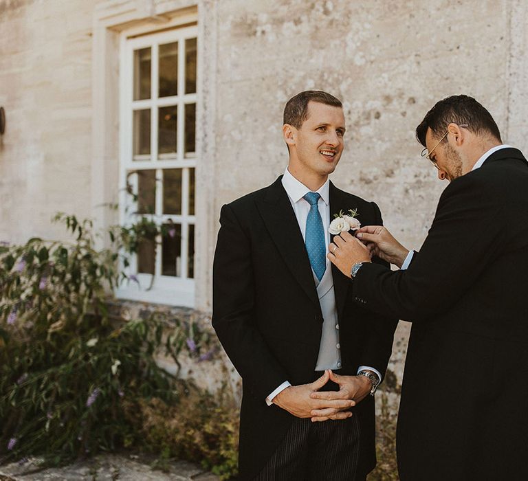 Groom in black morning suit with light grey waistcoat and light blue tie gets his white flower buttonhole attached