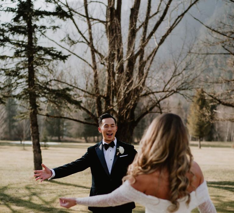 Bride in off shoulder sparkly mermaid wedding dress doing first look wedding photo with groom in classic black tuxedo, black bowtie and white garden rose boutonniere 