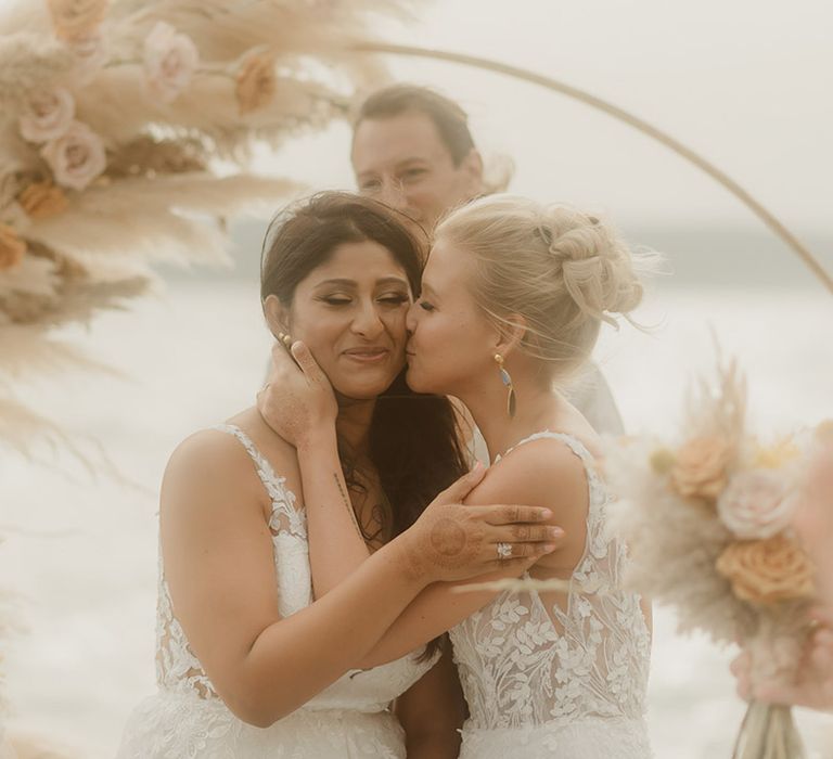 blonde bride with wedding chignon kissing her Indian bride at the altar during their beach wedding ceremony 