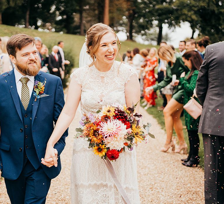 Bride in delicate short sleeve lace wedding dresss from Willowby by Watters with groom in blue suit walking out to confetti 