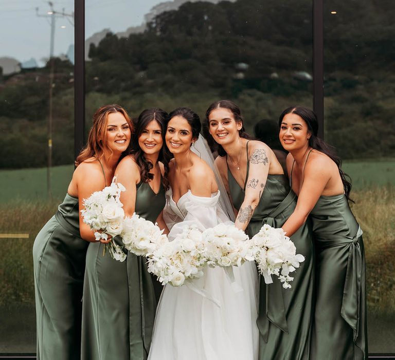 Bride in tulle wedding dress and bridesmaids in green satin. bridesmaid dresses lean forward with white wedding bouquets in front of Crumplebury wedding venue