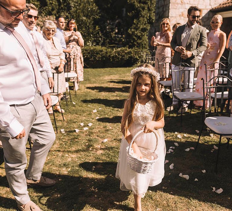 Flower girl in white dress wearing flower crown throwing petals out of wicker basket walking down the aisle 