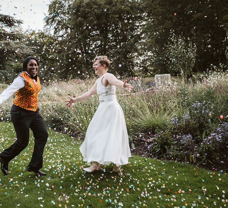 Bride in Ghanian patterned waistcoat with the bride in sleeveless wedding dress playing with pastel confetti 