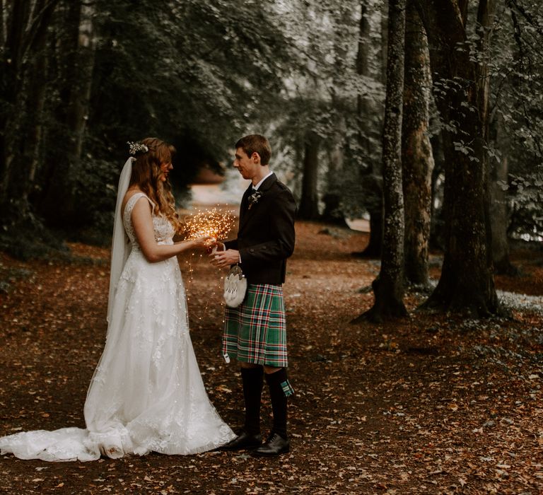 Bride in floral detailed wedding dress and groom in traditional kilt stand with fairy lights between them in the Scottish Highlands