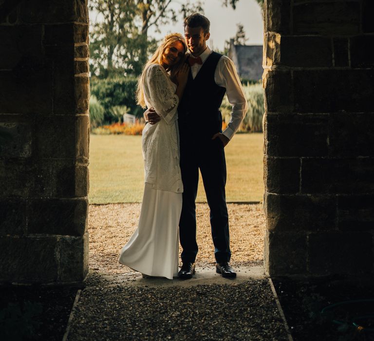 Bride and groom stand together under archway with bride in lace boho cover and groom with bright orange bow tie