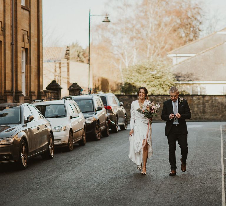 Brown haired bride walks with father carrying pastel bouquet tied with pink ribbon 