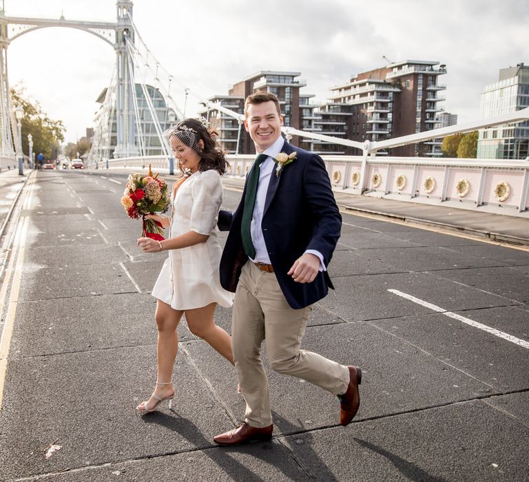 Groom in beige trousers and navy jacket crossing the road with his bride in a short wedding dress and birdcage veil