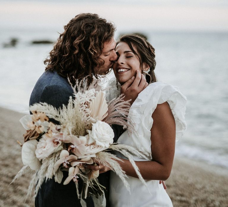 Groom with curly hair kissing his brides cheek on the beach whilst she holds her orchid wedding bouquet with anthuriums and pampas grass