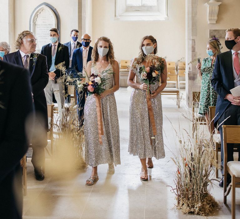 Bridesmaids in silver sequin dresses holding wildflower wedding bouquets tied with ribbon walking down the aisle 