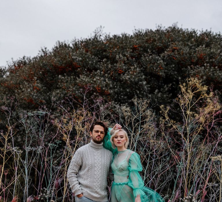 Stylish bride in an all in one body suit with a sheer pastel wedding dress standing neck to her groom on the beach in a grey jumper and jeans 