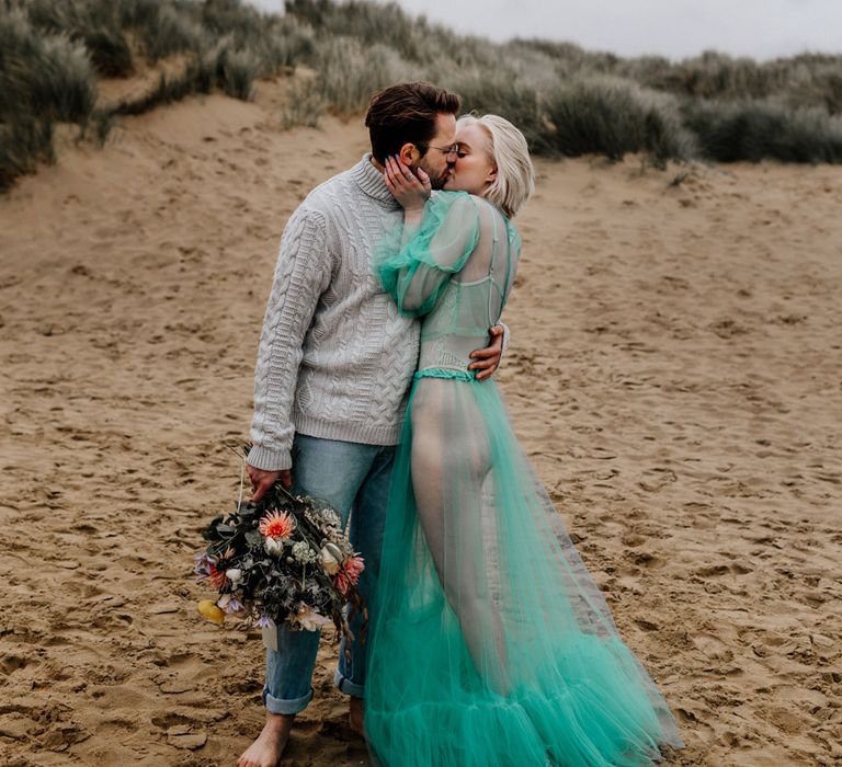 White bearded groom in a grey jumper and rolled up stone wash jeans kissing his bride in a sheer pastel wedding dress on the beach at their elopement 