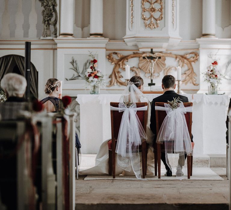 Bride and groom sit at the altar on burgundy chairs with white tulle details waiting to be married