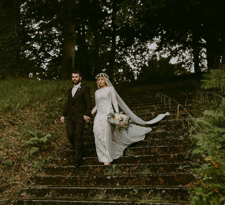 Bride in Grace Loves Lace wedding dress and single tier cathedral length veil walks through forest holding hands with groom in black Hugo Boss suit at Drenagh Estate Wedding