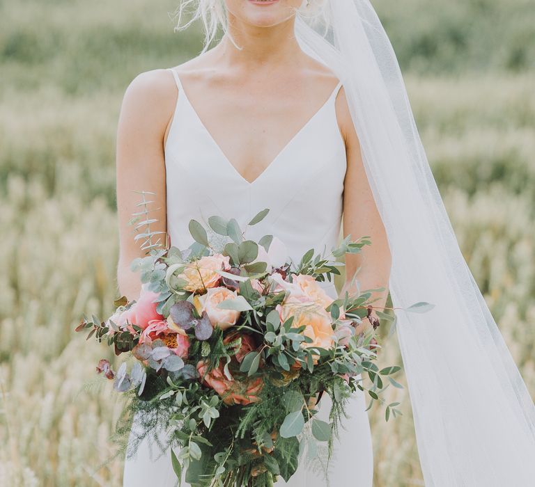 Bride smiles at the camera with her blonde hair pulled back into a low up-do and carrying pastel bouquet