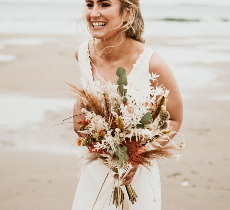 Bride laughs on the beach as the wind blows her hair and she carries Autumnal bouquet