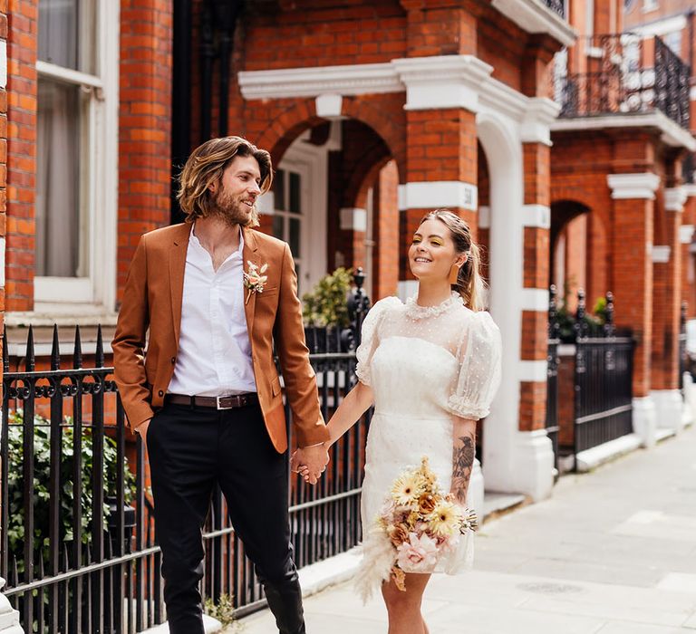 Groom in black trousers, white shirt and rust blazer holding hands with his bride in a polka dot short wedding dress