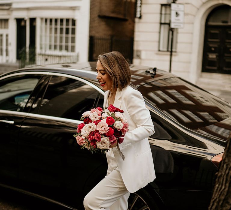 Bride walks to wedding ceremony in bridal jumpsuit whilst holding red and pink floral bouquet