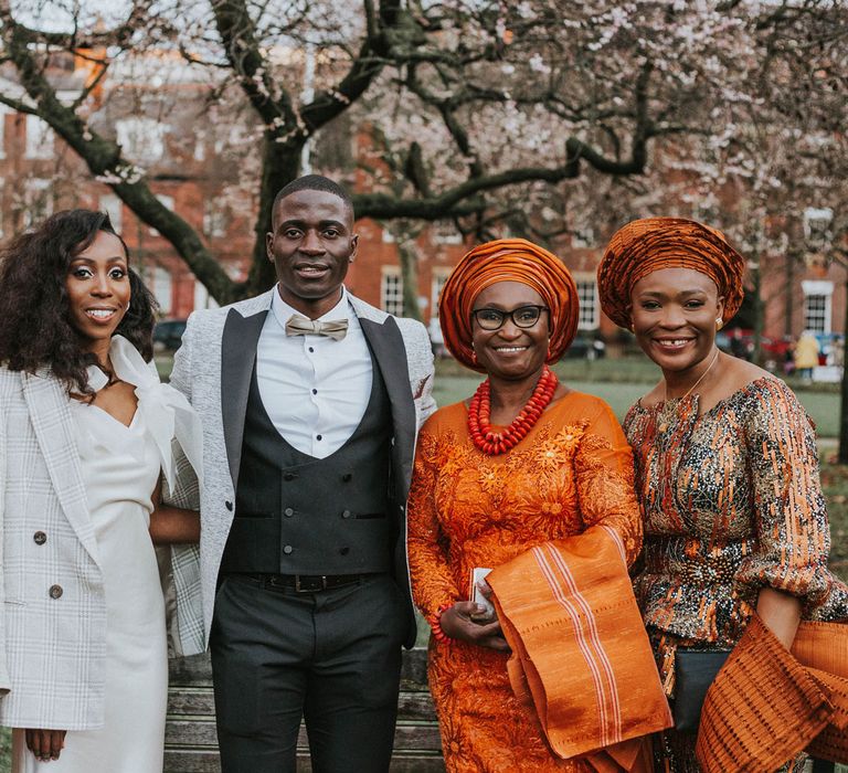 Bride in white Bec + Bride satin wedding dress and grey checked blazer stands with groom in grey suit jacket with black waistcoat and trousers and three wedding guests in traditional orange and white African outfits