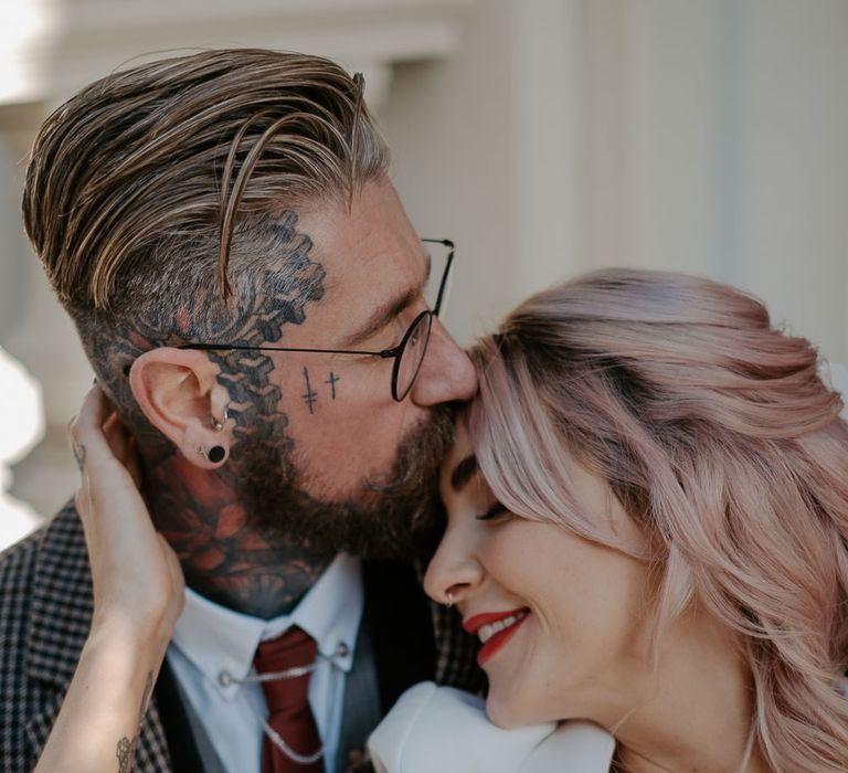 Tattooed groom kisses bride with pink blonde hair on head