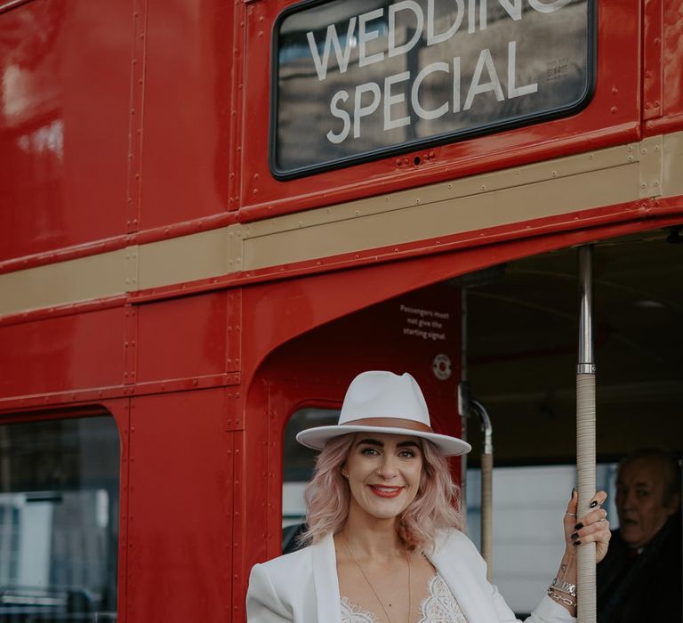 White bride in white suit with lace top and white fedora holds on to the pole of a red London bus at Marylebone wedding