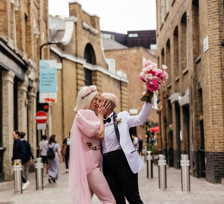 Bride in a pink jumpsuit kissing her bride in a white tuxedo jacket 