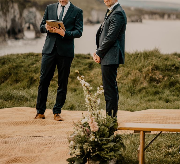 Male wedding celebrant in dark blue suit stands next to groom in dark blue suit waiting for bride on clifftop at Dunluce Castle wedding