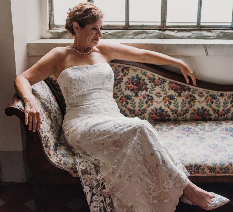 Bride sits on Chaise Longue whilst wearing an embroidered wedding gown with diamond necklace 