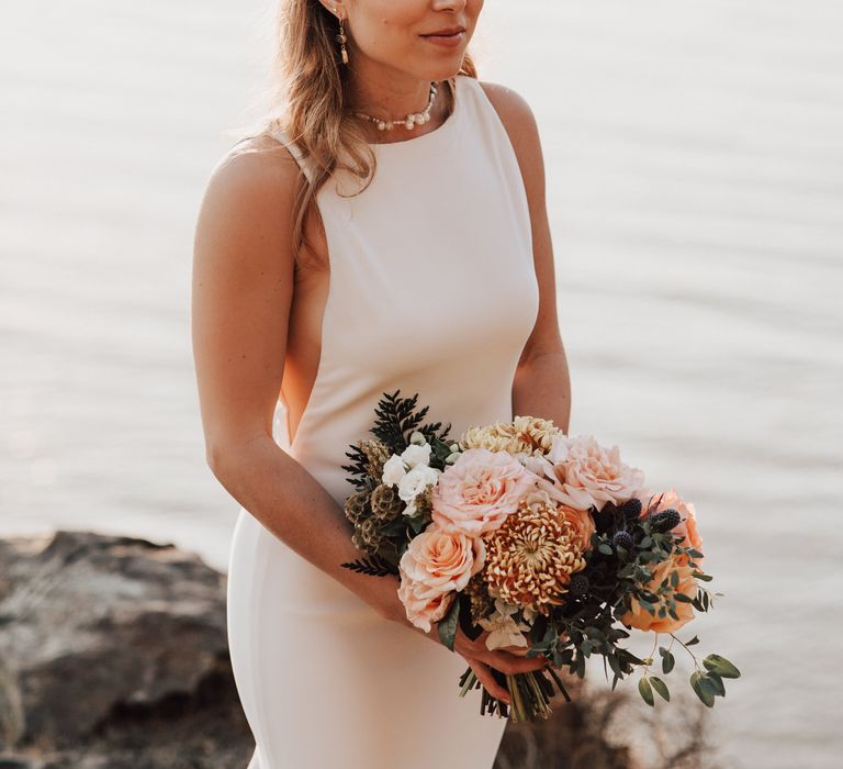 Bride stands in front of the ocean on the cliffside whilst the sun sets and she holds peach coloured floral bouquet with green foliage