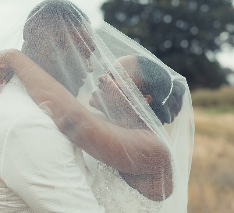 Black bride & groom stand beneath brides veil outdoors as she wraps her arms around his shoulders